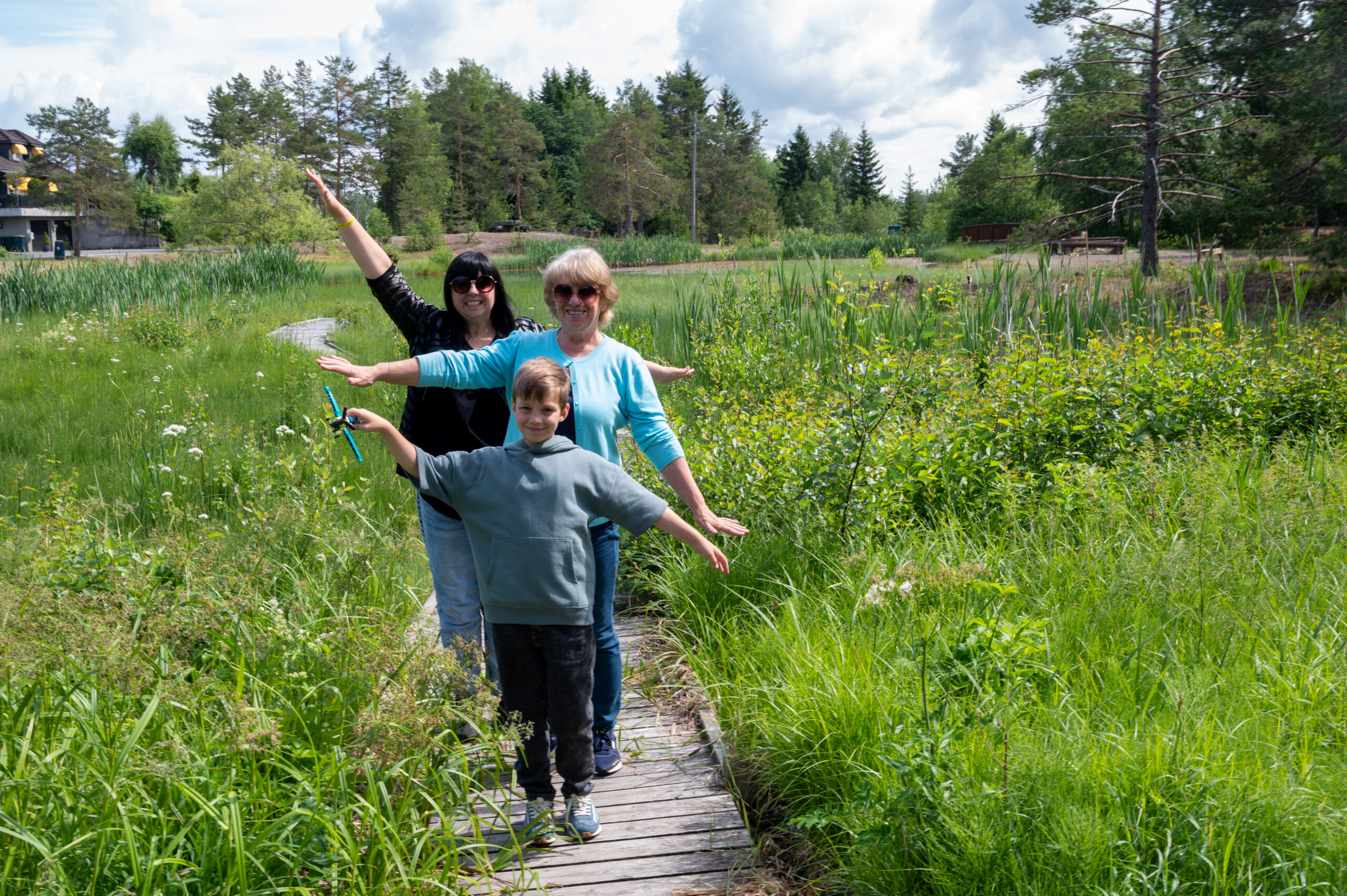 En gutt, en mor og en bestemor står ute på en sti. Rundt er det høyt gress. De strekker hendene ut til siden.