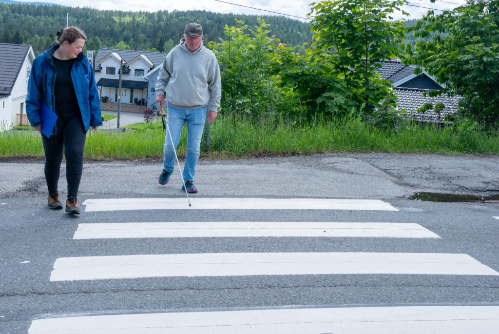 To personer går over et gangfelt. Den ene benytter mobilitetsstokk