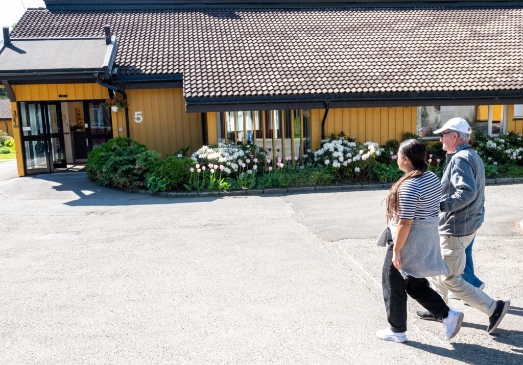 To personer går mot en inngang. Ved siden av inngangen er det lave busker og blomster