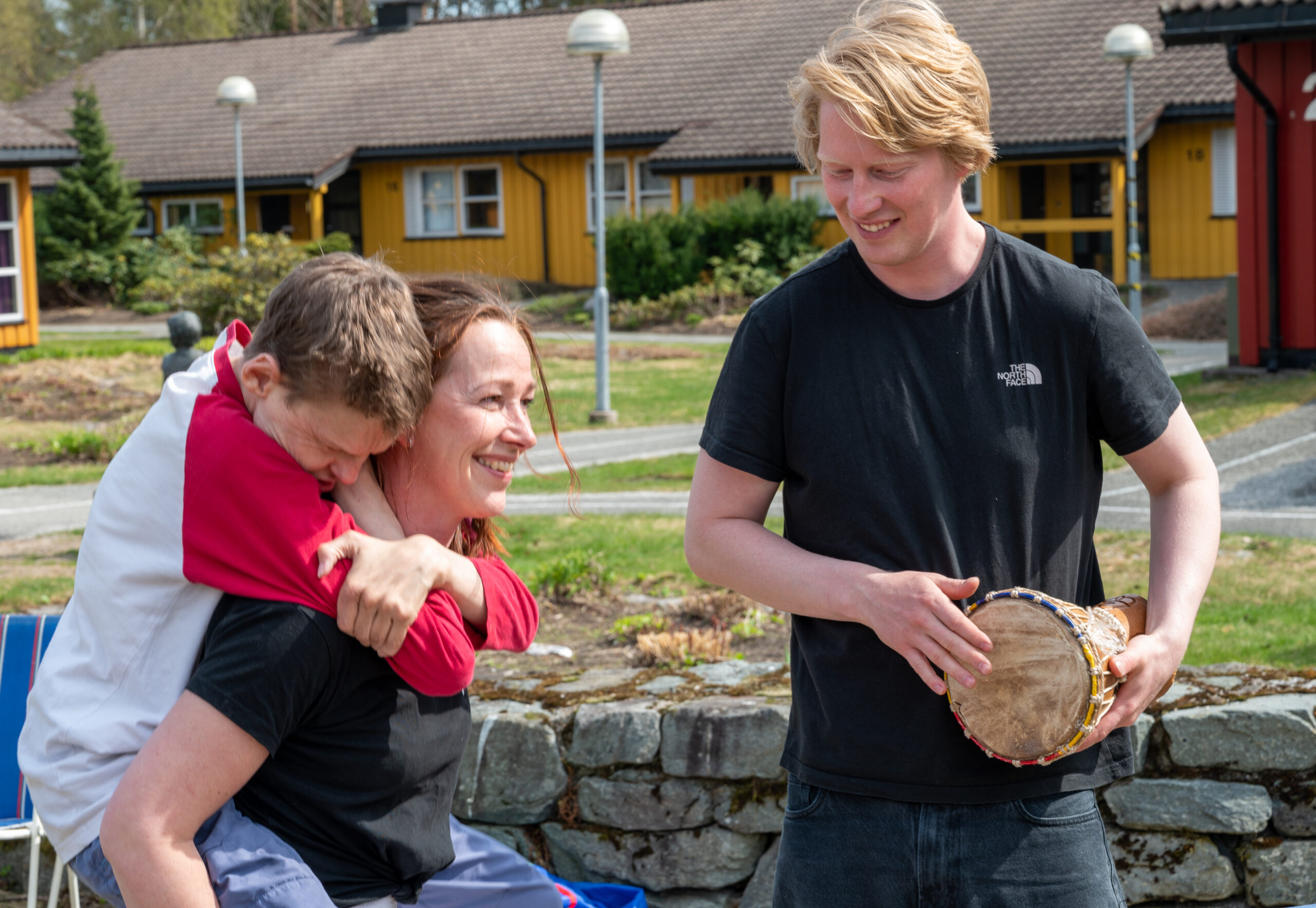 En person på ryggen til en annen. Hen holder armene godt rundt halsen og legger hodet inntil. En person står ved siden av med en tromme.