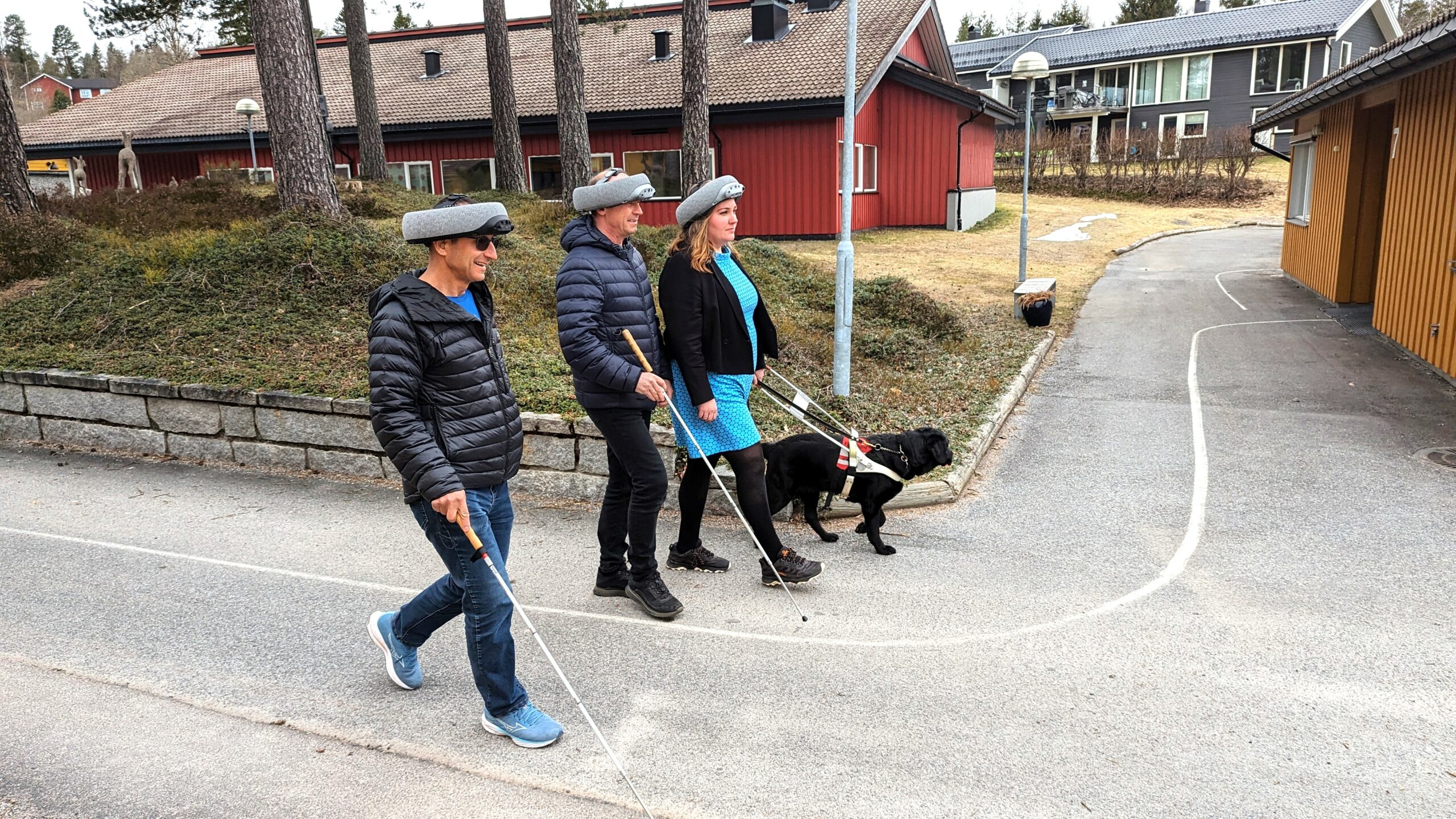 Tre personer går ute med superbrain hjelmer på hodet. To bruker mobilitetsstokk og en førerhund.