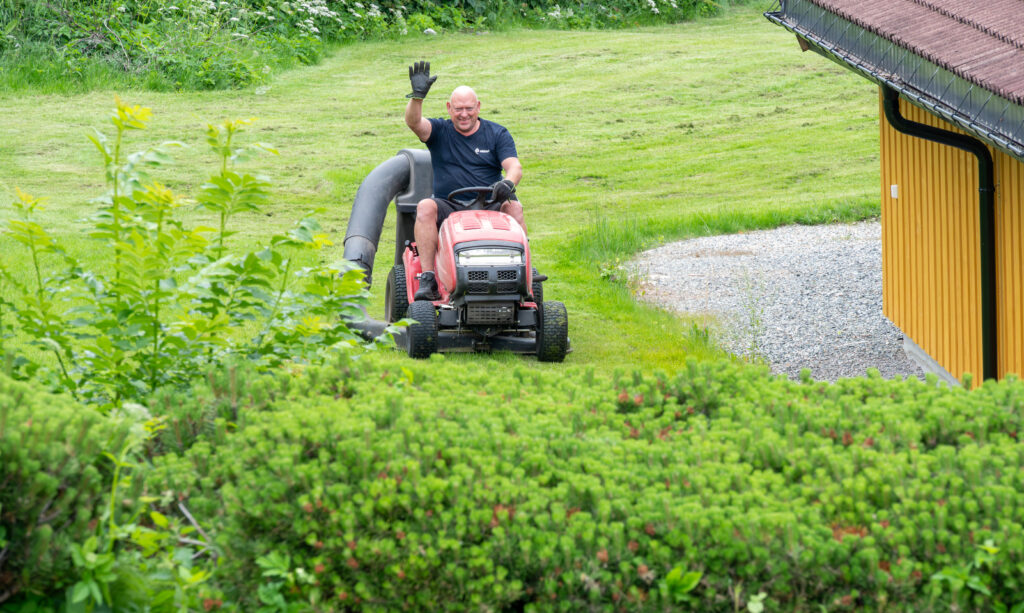 Vaktmester sitter på en plenklippertraktor. Rundt vises gress og busker.